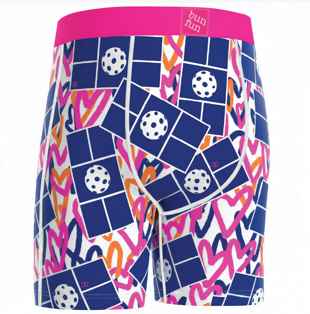 Men's boxer briefs featuring a vibrant abstract pattern of navy blue blocks, pink scribbles, orange zig-zags, and white soccer balls on a white background, with a bright pink waistband.
