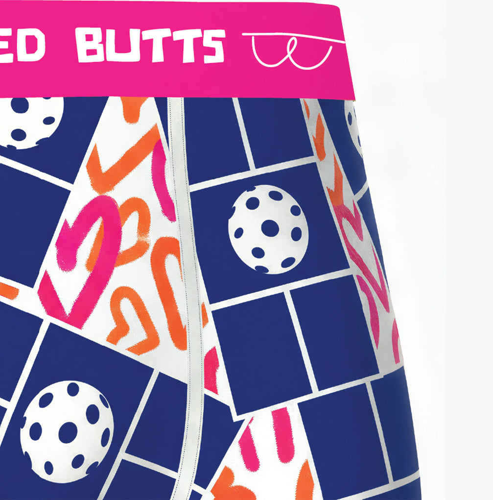 Close-up of patterned boxer briefs with a bright pink waistband reading 'PICKLED BUTTS' and white line art of glasses. The fabric has a navy grid background with white, pink, and orange abstract shapes and polka-dot circle motifs.