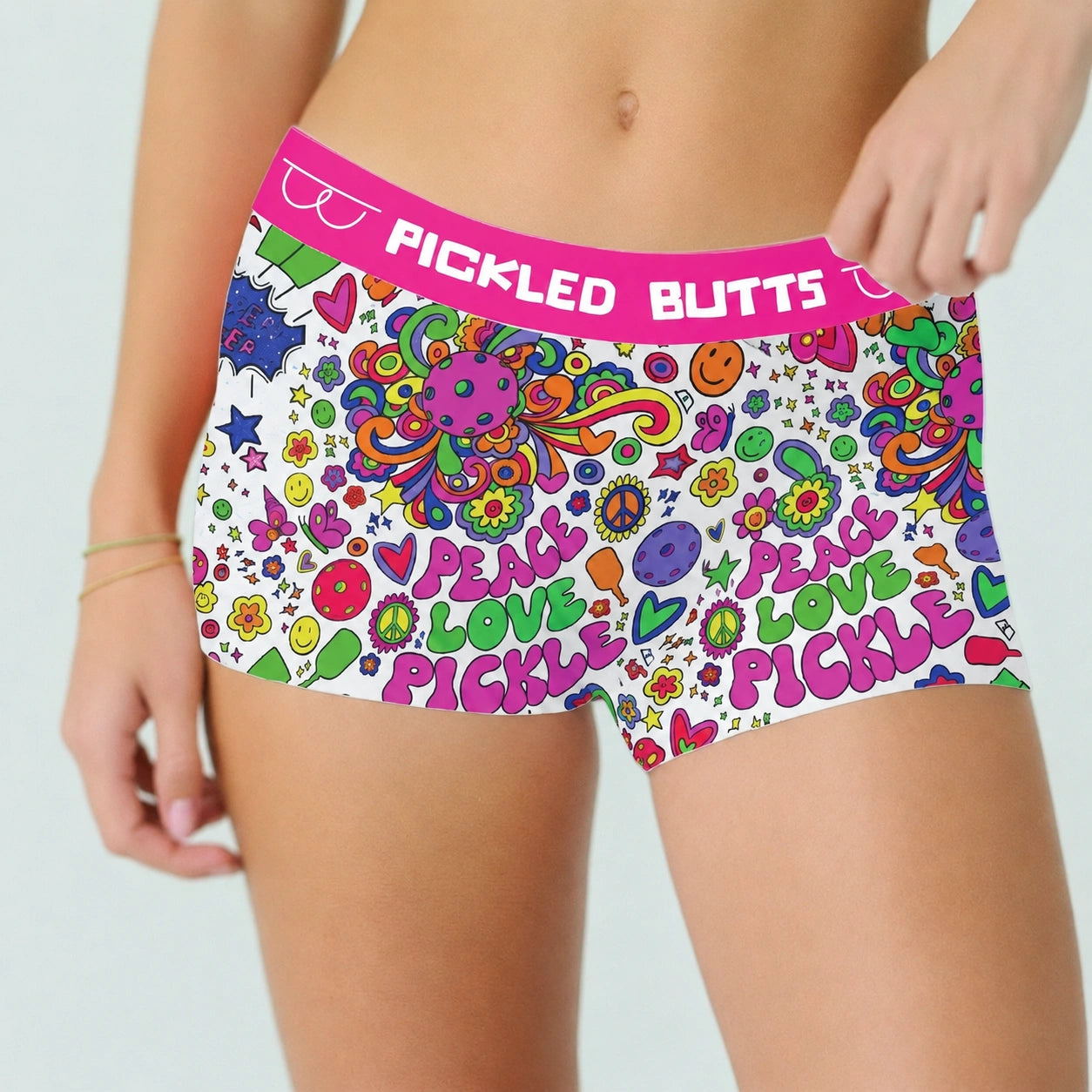 Women wearing Hippy Pickleball Underwear for Women with a bright pink waistband and a colorful retro doodle print. The underwear has the words “PICKLED BUTTS” across the waistband and repeated “PEACE LOVE PICKLE” text and fruit/abstract motifs across the front.