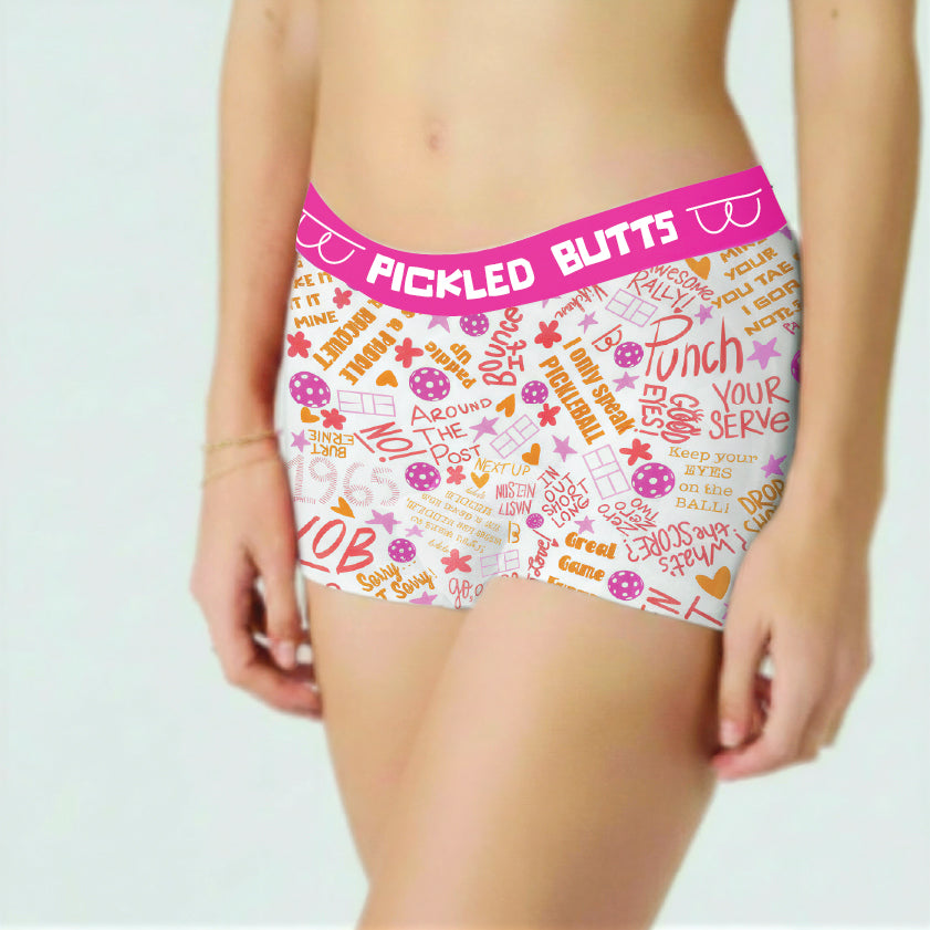 White women's boxer briefs with a bright pink waistband and a colorful retro-themed graphic print featuring text like 'PICKLED BUTTS' and 'PUNCH YOUR SERVE'. The underwear is modeled on a person against a plain white background.